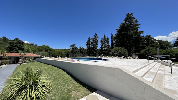 A serene outdoor setting featuring a large, tiled pool area with several lounge chairs neatly aligned along the pool. The surrounding landscape is lush with green grass and tall trees. A small building with a red-tiled roof is visible to the left, and the sky is clear with deep blue colors.