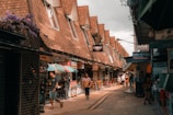 A bustling street scene with people walking through a narrow lane lined with small shops. The shops have slanted roofs and display various signs. Umbrellas are open over some of the stalls, adding a splash of color. The atmosphere is lively with both locals and tourists exploring the area.