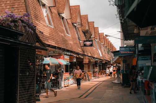 A bustling street scene with people walking through a narrow lane lined with small shops. The shops have slanted roofs and display various signs. Umbrellas are open over some of the stalls, adding a splash of color. The atmosphere is lively with both locals and tourists exploring the area.