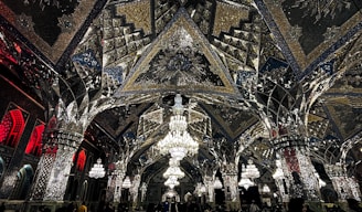 An elaborate interior featuring intricate mirrored ceilings and ornate chandeliers. The design includes geometric patterns, reflective surfaces, and detailed artwork. Warm lighting enhances the glass and metallic textures, creating a luxurious and sophisticated ambiance.