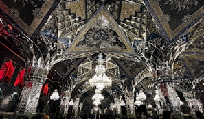 An elaborate interior featuring intricate mirrored ceilings and ornate chandeliers. The design includes geometric patterns, reflective surfaces, and detailed artwork. Warm lighting enhances the glass and metallic textures, creating a luxurious and sophisticated ambiance.