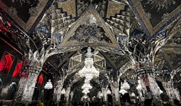 An elaborate interior featuring intricate mirrored ceilings and ornate chandeliers. The design includes geometric patterns, reflective surfaces, and detailed artwork. Warm lighting enhances the glass and metallic textures, creating a luxurious and sophisticated ambiance.