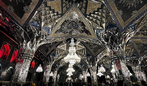 An elaborate interior featuring intricate mirrored ceilings and ornate chandeliers. The design includes geometric patterns, reflective surfaces, and detailed artwork. Warm lighting enhances the glass and metallic textures, creating a luxurious and sophisticated ambiance.