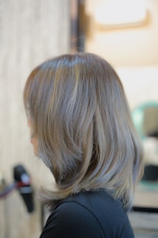A person with light brown hair, featuring shades of ash and silver, styled in a smooth and slightly layered cut seen from the side. The background is softly blurred, suggesting an indoor environment possibly in a salon.