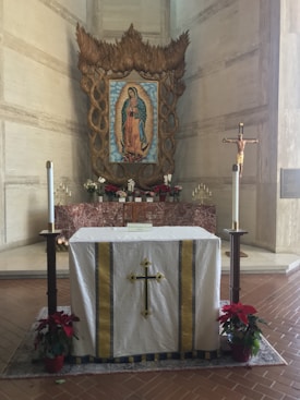 An altar adorned with a white cloth featuring a central yellow cross. The background includes a large framed image of a religious figure surrounded by intricate wood carvings. On either side of the altar are two tall candles, and potted red poinsettia plants are placed at the base.