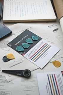 A collection of design and technical documents lay scattered on a desk. A set of guitar picks, each in different colors such as teal, yellow, and black, is prominently displayed. There is a smartphone partially visible and various papers with technical drawings and notes. A measuring tool resembling a compass is also on the table, adding to the theme of design and planning.