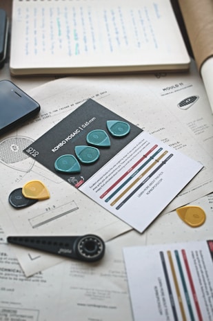 A collection of design and technical documents lay scattered on a desk. A set of guitar picks, each in different colors such as teal, yellow, and black, is prominently displayed. There is a smartphone partially visible and various papers with technical drawings and notes. A measuring tool resembling a compass is also on the table, adding to the theme of design and planning.