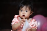 A young child with dark hair wearing a pink, pearl-adorned dress is holding a piece of paper and looking curiously at the camera. The background features pink balloons, contributing to a festive setting.