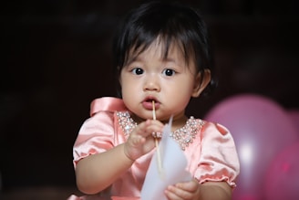 A young child with dark hair wearing a pink, pearl-adorned dress is holding a piece of paper and looking curiously at the camera. The background features pink balloons, contributing to a festive setting.
