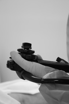 A gloved hand operating a medical endoscope against a neutral background. The focus is on the handle, which has control buttons and knobs.