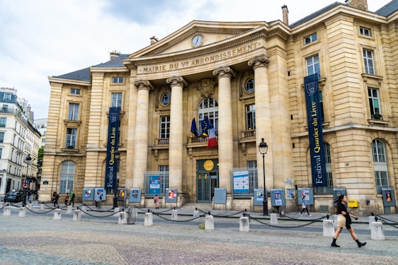 A grand historic building with neoclassical architecture featuring large columns and ornate stonework. There are flags flying above the entrance and banners advertising a book festival hanging from the facade. A cobblestone square is in front of the building, with people walking by.