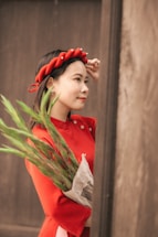 A woman wearing a vibrant red dress and a braided headband holds a bunch of green leaves wrapped in newspaper. She is gazing forward and slightly smiling, standing near a wooden wall.