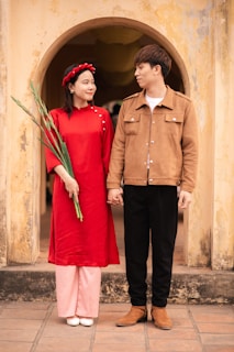 A couple stands side by side in front of an aged, arched doorway. The woman wears a bright red traditional dress with a matching headband and holds long green leaves. The man is dressed in a brown jacket, black pants, and brown boots. They are holding hands and looking at each other affectionately.
