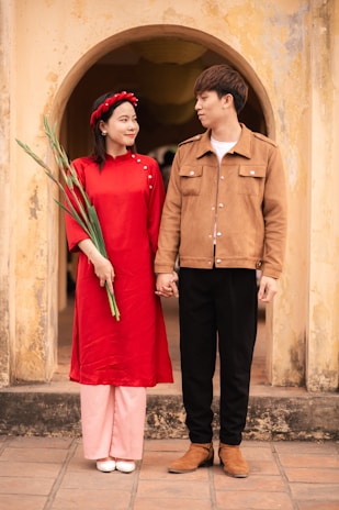 A couple stands side by side in front of an aged, arched doorway. The woman wears a bright red traditional dress with a matching headband and holds long green leaves. The man is dressed in a brown jacket, black pants, and brown boots. They are holding hands and looking at each other affectionately.