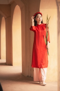 A woman wearing a traditional red dress and a matching headpiece holds a green plant while standing in an architectural setting with tall arches and warm lighting.
