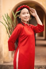A woman is dressed in a vibrant red traditional outfit, holding a bundle of green plants. She is smiling warmly and wearing a matching red headdress, standing against an arched, neutral-colored background.