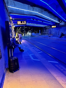 A dimly lit tram station with an overhead structure illuminated by bright blue lights. Several people are waiting near the tram tracks, with one person having a suitcase next to them. The station is mostly empty, with signage displaying information in the background.