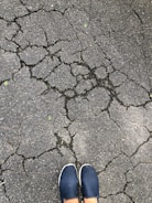 Cracks spread across a grey asphalt surface, forming an intricate network. At the bottom of the image, a pair of feet in blue slip-on shoes are visible.