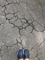Cracks spread across a grey asphalt surface, forming an intricate network. At the bottom of the image, a pair of feet in blue slip-on shoes are visible.