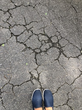 Cracks spread across a grey asphalt surface, forming an intricate network. At the bottom of the image, a pair of feet in blue slip-on shoes are visible.