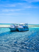 Two white yachts are anchored close together on a bright blue sea under a clear sky. The water is sparkling in the sunlight, creating a vibrant, inviting scene. There are a few people visible on the yachts, enjoying the serene environment.