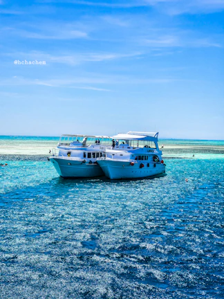 Two white yachts are anchored close together on a bright blue sea under a clear sky. The water is sparkling in the sunlight, creating a vibrant, inviting scene. There are a few people visible on the yachts, enjoying the serene environment.