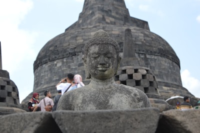 A stone statue of Buddha is prominently featured in the foreground, partially surrounded by ancient stupa structures. The large, layered stone dome of another stupa is visible behind the statue. Several people, some wearing head coverings, are gathered in the background, indicating a tourist spot or cultural site.