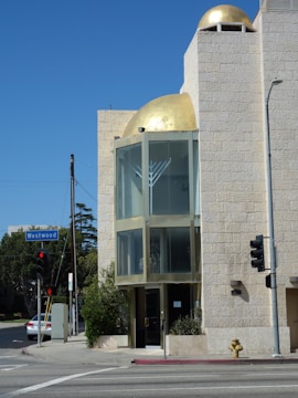 An urban street scene features a modern building with beige stone walls and a prominent golden dome at the top. There is an intersection with a traffic light, a hydrant, and a street sign labeled 'Westwood'. A menorah symbol is visible on the glass facade of the building.