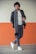 A man is leaning against a wall with one hand in his pocket and the other adjusting his scarf. He is dressed in a casual yet stylish outfit that includes a leather jacket, a white t-shirt, gray trousers, a gray scarf with black ends, and white sneakers. The background wall has a two-tone color with the upper part in light beige and the lower part in orange.