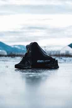 A pair of black sports shoes and a black bag with the brand name 'Bauer' are placed on an icy surface. The background features blurred mountains and a cloudy sky, creating a serene and somewhat cold atmosphere.
