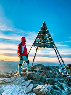 A person dressed in hiking gear stands on a rocky mountainous peak near a metal pyramid structure. The sky is clear with a few clouds, and the sun is setting or rising, casting a warm glow over the scene.