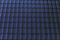 Dark, glossy roof tiles are arranged in a neat and uniform pattern. The tiles have a wavy design with a reflective surface, showing some minor stains or imperfections.