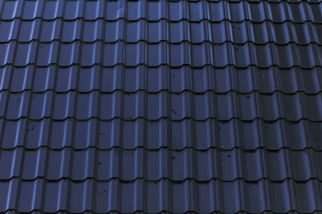 Dark, glossy roof tiles are arranged in a neat and uniform pattern. The tiles have a wavy design with a reflective surface, showing some minor stains or imperfections.