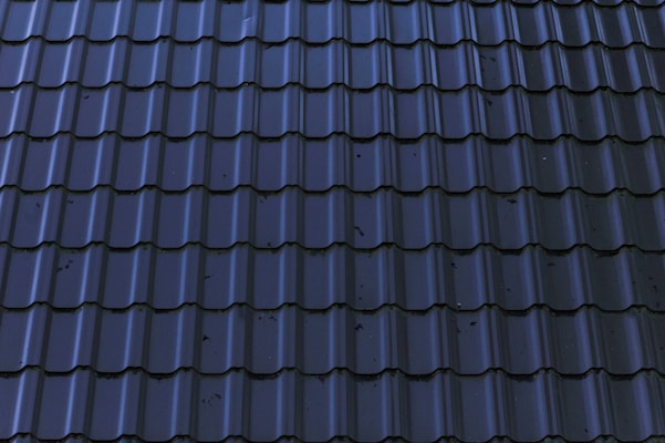 Dark, glossy roof tiles are arranged in a neat and uniform pattern. The tiles have a wavy design with a reflective surface, showing some minor stains or imperfections.