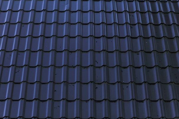 Dark, glossy roof tiles are arranged in a neat and uniform pattern. The tiles have a wavy design with a reflective surface, showing some minor stains or imperfections.