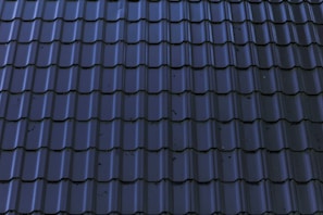 Dark, glossy roof tiles are arranged in a neat and uniform pattern. The tiles have a wavy design with a reflective surface, showing some minor stains or imperfections.