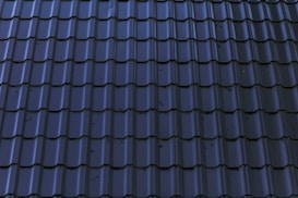 Dark, glossy roof tiles are arranged in a neat and uniform pattern. The tiles have a wavy design with a reflective surface, showing some minor stains or imperfections.
