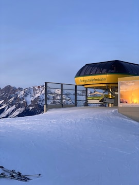 A ski lift station sits atop a snowy mountain slope, with tracks left by skis visible in the snow. The station has a modern design with a dark curved roof and yellow accents. Majestic snow-covered peaks are seen in the background under a clear blue sky, and a set of skis rests in the snow.