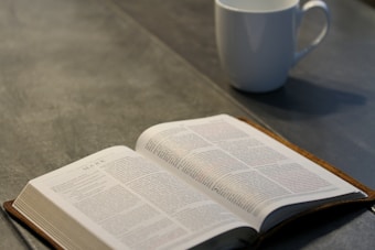 An open book is displayed on a flat surface, likely a table, with readable text. To the right of the book, a white ceramic mug is placed, likely containing a beverage. The setting is well-lit, creating a calm and serene atmosphere.