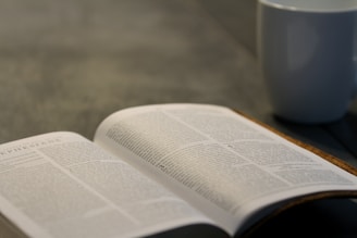 An open book with visible text, possibly a Bible, featuring a page from Ephesians. A white ceramic mug is positioned in the background on a smooth, dark surface.