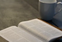 An open book lies on a table with text visible from a religious scripture, accompanied by a white ceramic mug. The setting is calm and suggests a moment of quiet study or reflection.
