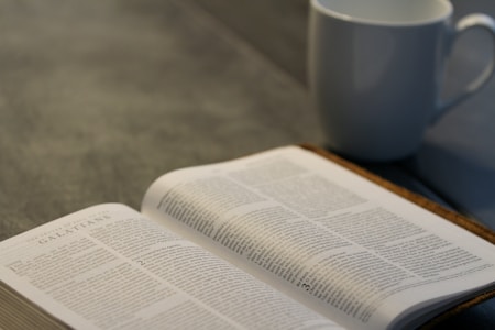 An open book lies on a table with text visible from a religious scripture, accompanied by a white ceramic mug. The setting is calm and suggests a moment of quiet study or reflection.