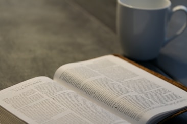 An open book lies on a table with text visible from a religious scripture, accompanied by a white ceramic mug. The setting is calm and suggests a moment of quiet study or reflection.