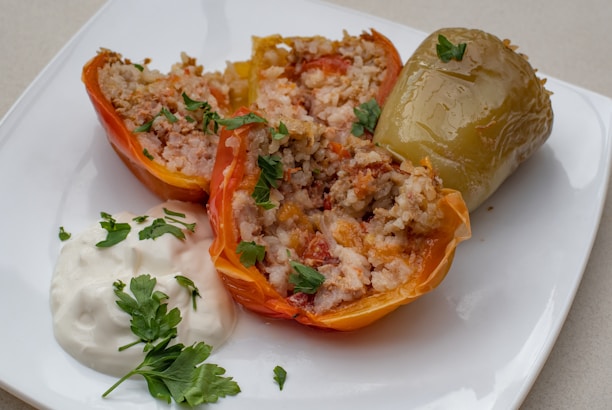 A plate featuring stuffed peppers filled with a mixture of rice, ground meat, and vegetables. One pepper is cut in half to reveal the filling, while the other is whole. The dish is garnished with fresh parsley and served alongside a dollop of sour cream.
