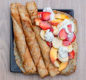 A plate of crepes is presented, featuring three rolled crepes and one folded crepe topped with sliced strawberries, bananas, and peaches. Whipped cream is dolloped on top, adding a fresh and appetizing touch. The plate sits on a light wooden surface, enhancing the vibrant colors of the fruit.
