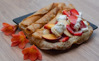A plate of crepes adorned with slices of strawberries, bananas, and peaches, topped with dollops of whipped cream. The crepes have a golden-brown hue and are accompanied by vibrant orange flowers on a light wooden surface.