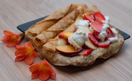 A plate of crepes adorned with slices of strawberries, bananas, and peaches, topped with dollops of whipped cream. The crepes have a golden-brown hue and are accompanied by vibrant orange flowers on a light wooden surface.