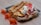 A plate of crepes adorned with slices of strawberries, bananas, and peaches, topped with dollops of whipped cream. The crepes have a golden-brown hue and are accompanied by vibrant orange flowers on a light wooden surface.