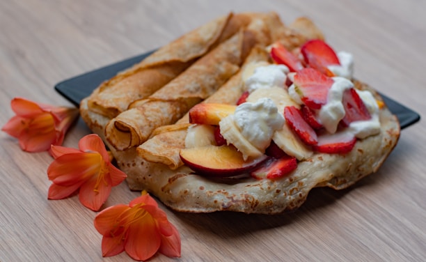 A plate of crepes adorned with slices of strawberries, bananas, and peaches, topped with dollops of whipped cream. The crepes have a golden-brown hue and are accompanied by vibrant orange flowers on a light wooden surface.