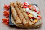Three rolled crepes are placed on a square black plate with an additional unfolded crepe beside them. The unfolded crepe is topped with slices of strawberries, bananas, and peaches, along with dollops of whipped cream. Bright orange flowers artfully decorate the backdrop on the wooden table surface.